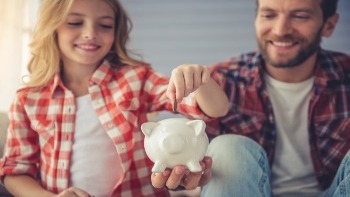 A father holds out a piggy bank so his daughter can place a quarter inside. 