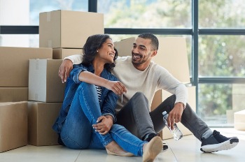 A young couple smile at each other as they sit and rest surrounded by moving boxes in their new home. 