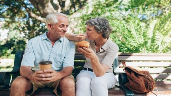 Elderly couple enjoy coffee and a muffin while sitting on a park bench on a sunny day. 